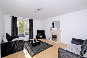 A modern living room with a fireplace, black and grey sofas, a flat-screen TV, and a large window with curtains. - Orange Apartments Beechgrove (Aberdeen)