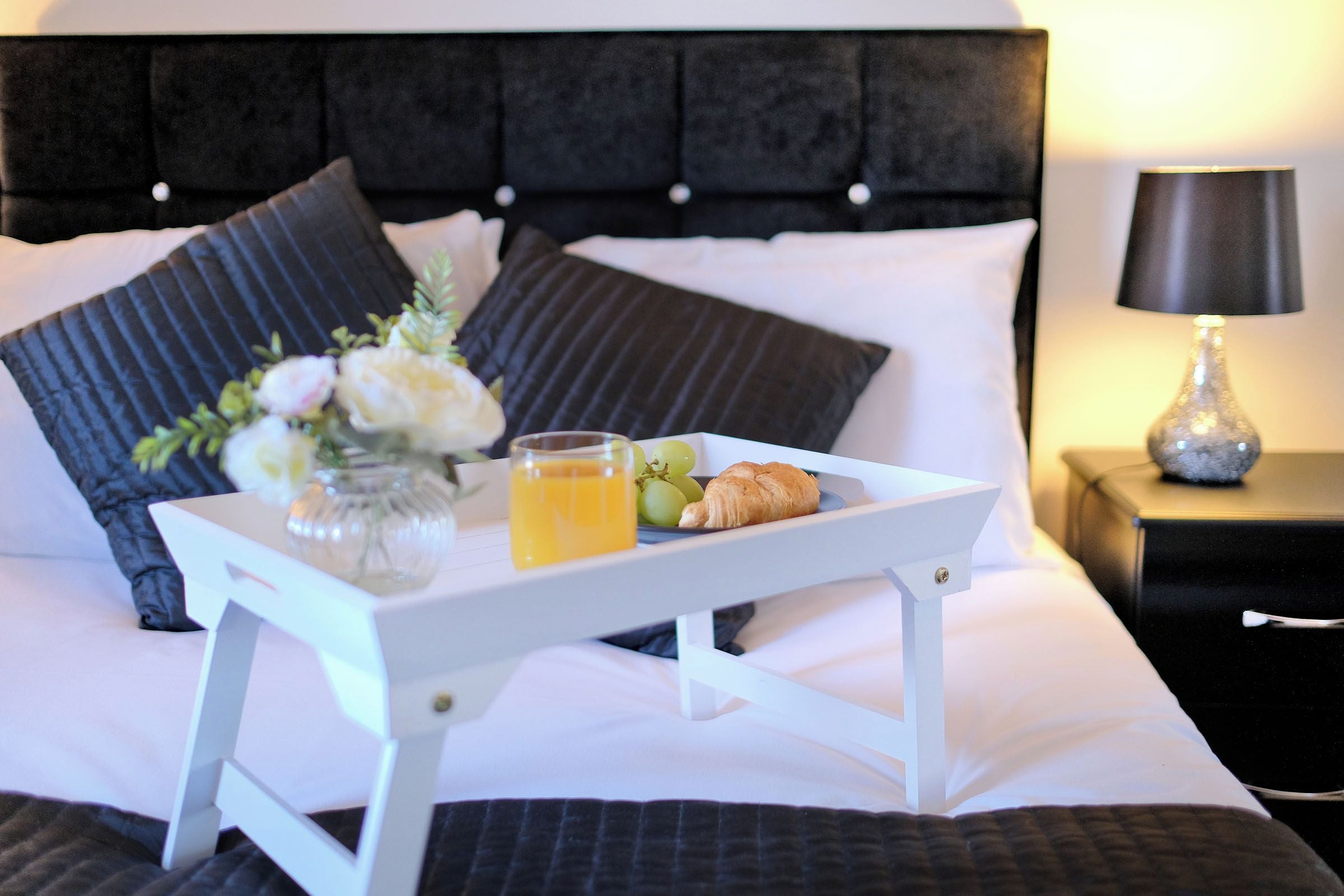 A neatly made bed with a tray of breakfast items including croissants, grapes, and orange juice, accompanied by a vase of flowers.