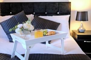 A neatly made bed with a tray of breakfast items including croissants, grapes, and orange juice, accompanied by a vase of flowers.