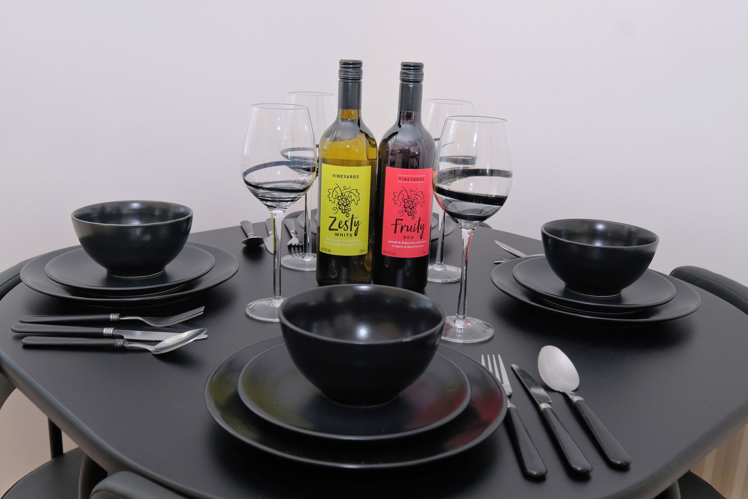 A table set with black bowls, wine glasses, and cutlery, with two bottles of wine labeled 'Zesty White' and 'Fruity Red'.