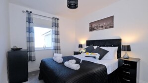 A hotel room with a bed, bedside tables, a window with curtains, a cityscape picture on the wall, and a ceiling light fixture. - Orange Apartments Beechgrove (Aberdeen)