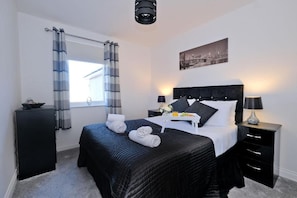 A hotel room with a bed, bedside tables, a window with curtains, a cityscape picture on the wall, and a ceiling light fixture. - Orange Apartments Beechgrove (Aberdeen)