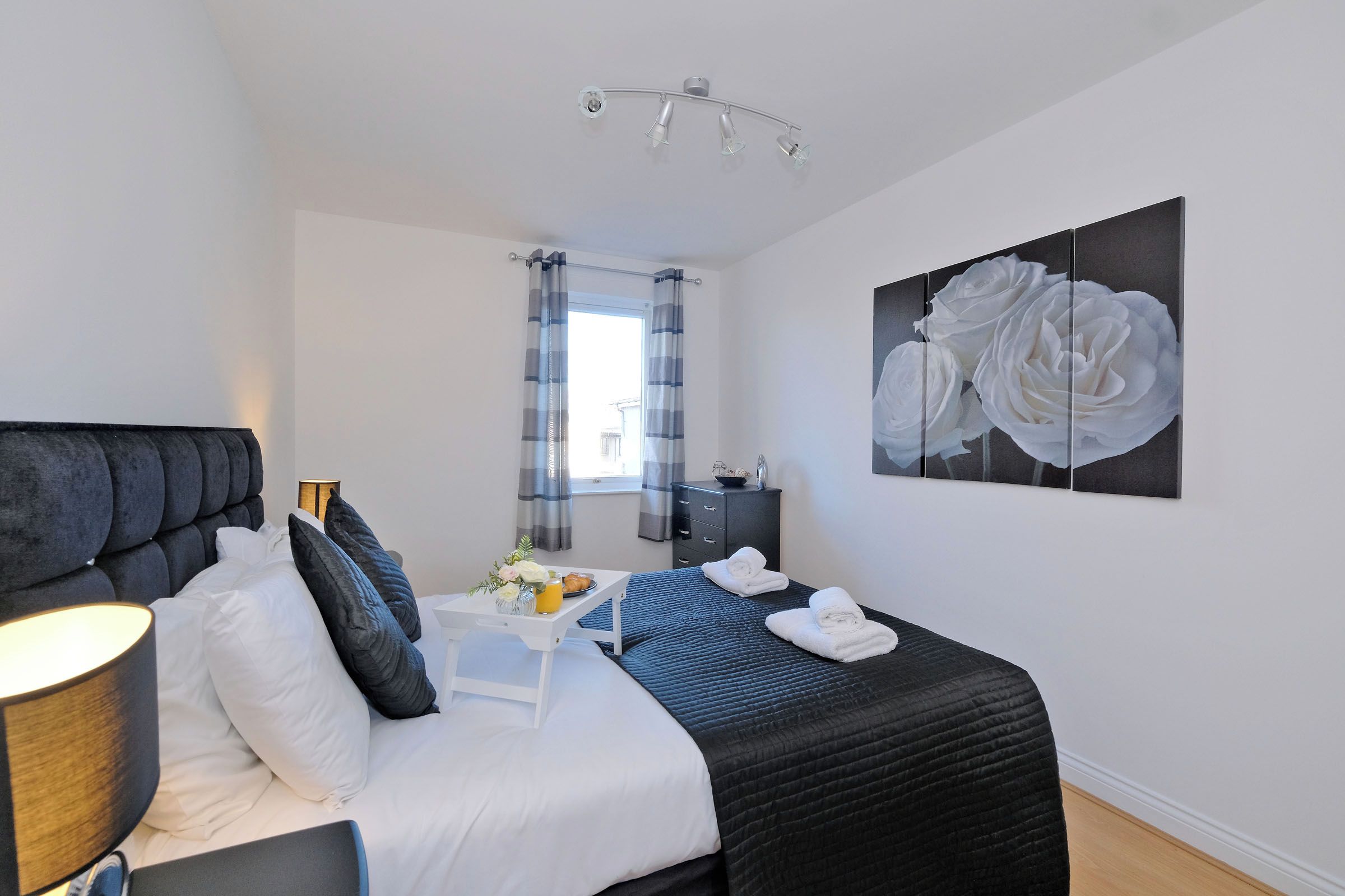 A hotel room with a bed, bedside table, lamp, and a painting of white roses.
