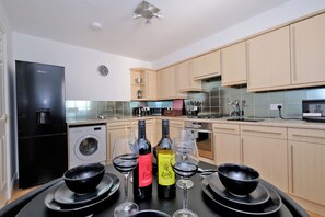 A modern kitchen with wooden cabinets, a black refrigerator, a washing machine, and a dining area with wine glasses and bottles. - Orange Apartments Beechgrove (Aberdeen)