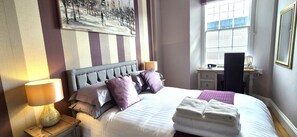 Een net opgemaakt bed met kussens, een nachtkastje met een lamp, een bureau met een stoel, en een raam met zonwering.