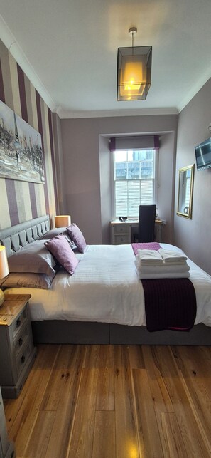 Een slaapkamer met een bed, nachtkastje, lamp, bureau, stoel, raam en kunst aan de muur.