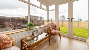 A spacious room with large windows, a wicker chair, a glass coffee table, and a view of a grassy field. - The Ferns (Ballymoney)