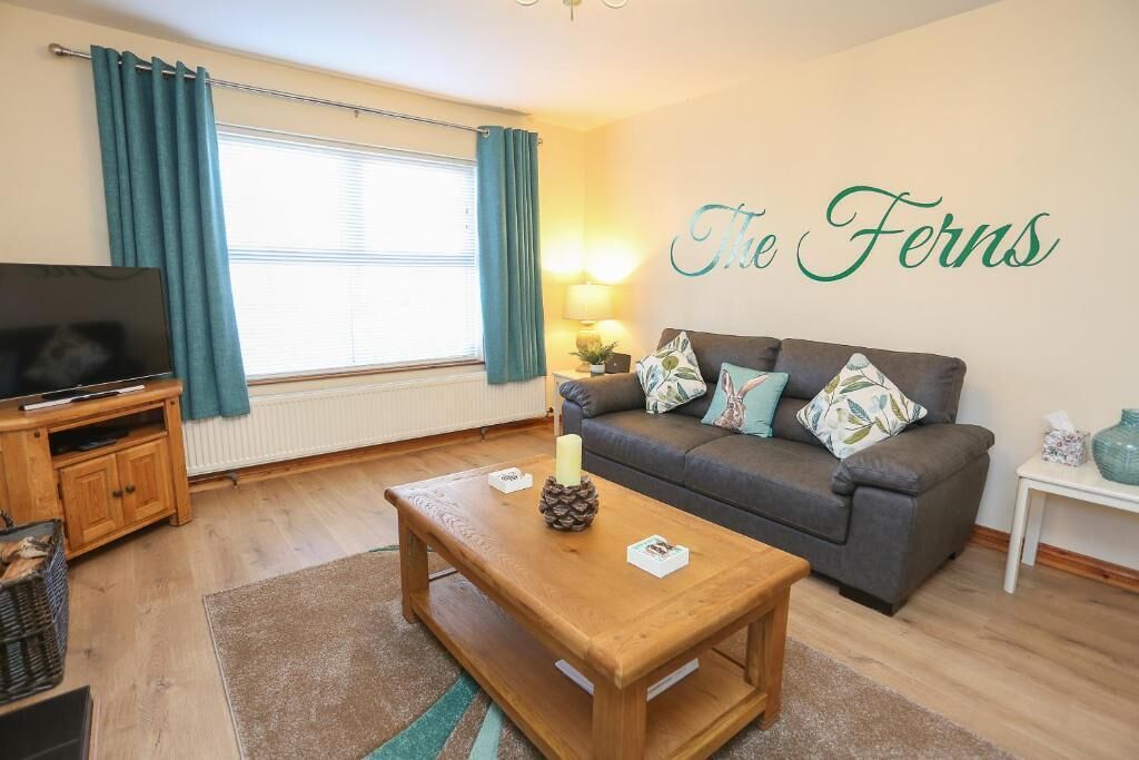 A living room with a sofa, a wooden coffee table, a television on a stand, and a wall with the text 'The Ferns'.