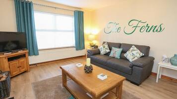 A living room with a sofa, a wooden coffee table, a television on a stand, and a wall with the text 'The Ferns'.