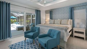 Quarto com uma cama grande, duas poltronas azuis, um ventilador de teto e vista para uma área de piscina externa.