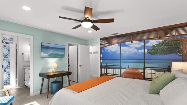 Quarto com cama, escrivaninha, cadeira, ventilador de teto e uma grande janela com vista para o oceano.