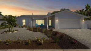 Uma casa moderna de dois andares com garagem, entrada por meio de um acesso de veículos e um jardim com paisagismo, diversas plantas e área com brita.