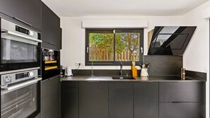 A modern kitchen with dark cabinetry, a built-in oven, a window with a view of trees, and a black range hood. - Villa Arasu, 10 pers, piscine chauffée, vue mer, 15 mn à pieds plage (STE LUCIE DE PORTO VECCHIO)