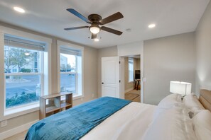 Un dormitorio con cama, ventilador de techo, mesita de noche con lámpara y amplias ventanas que dan a la calle.