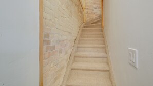 Una escalera estrecha con alfombra color crema y una pared de ladrillo a un lado.