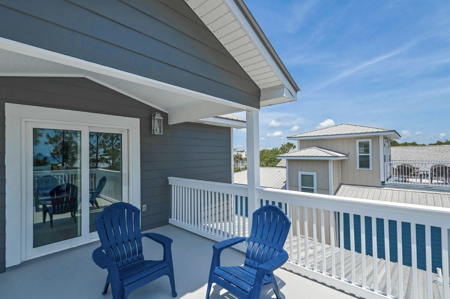 A balcony with two blue chairs, a glass door leading to a patio, and a view of neighboring houses.