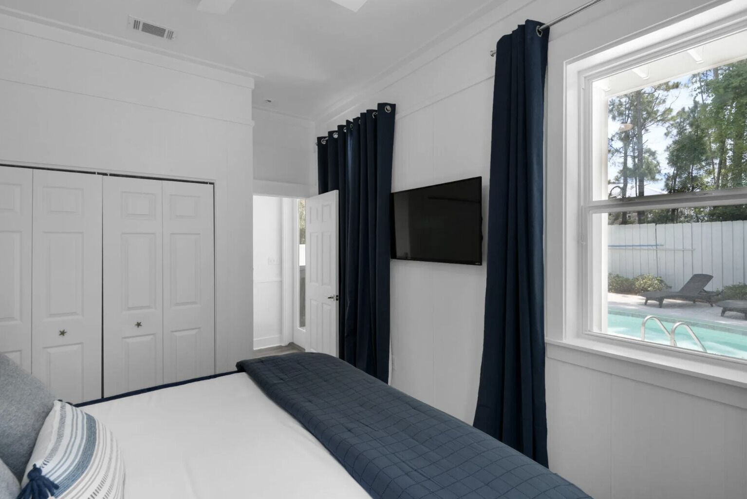 A bedroom with a bed, a TV, a window with curtains, and a view of a swimming pool.
