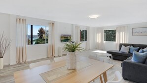 Un salon moderne comprenant un coin repas, de grandes fenêtres avec des rideaux, un canapé gris et une table basse blanche.
