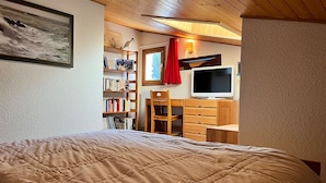 Un dormitorio con una cama, un aparador de madera, un televisor, una estantería y una ventana con cortina roja.