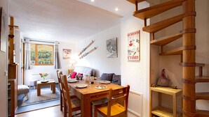 Un área de comedor con una mesa y sillas de madera, una mesita auxiliar con un jarrón de flores, y vista a un bosque a través de una ventana.