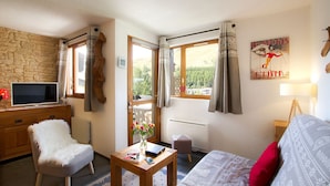Una habitación acogedora con una pared de piedra, un escritorio de madera con un televisor, una silla blanca, una mesita con un florero, un sofá con cojines estampados y una ventana con cortinas.