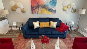 Un soggiorno con un divano blu scuro, un quadro astratto dai colori vivaci, elementi decorativi a parete e una coperta rossa.