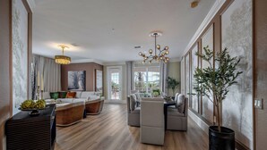 A spacious living room with a wooden floor, a dining area with a table and chairs, a large chandelier, and a potted plant.