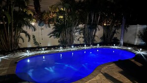 A swimming pool with a curved edge, surrounded by a brick patio and palm trees. - Peacock Paradise (Holmes Beach)