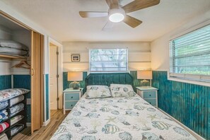 Un dormitorio con cama, ventilador de techo, ventana con persianas, mesita de noche con lámpara y armario con ropa colgada.