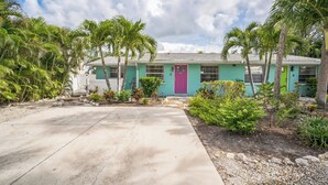 Une maison de plain-pied à la façade turquoise et à la porte violette, entourée de palmiers et bordée d’une allée en béton.