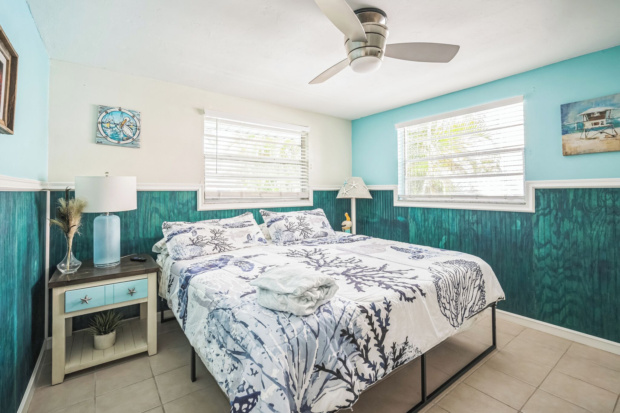A bedroom with a bed, two bedside tables, a ceiling fan, and two windows with blinds.