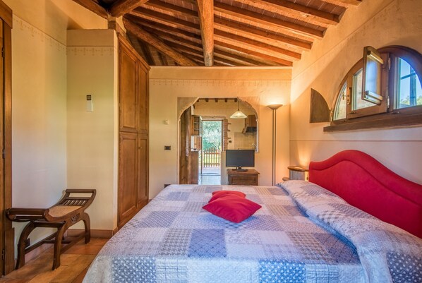 Une chambre avec un plafond en bois, un lit recouvert d’une couvre-lit à motifs, un oreiller rouge, un banc en bois et une porte donnant sur un balcon.