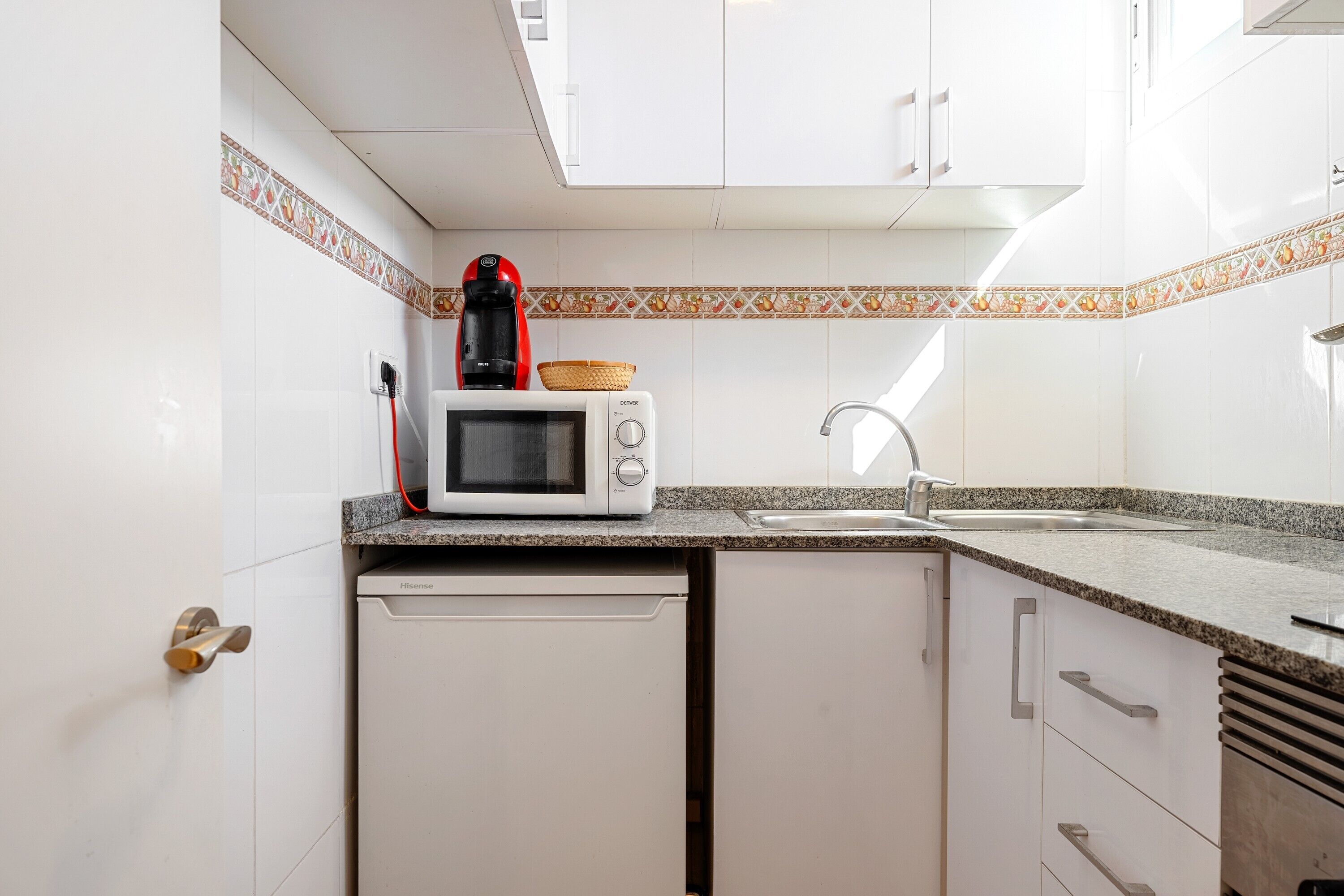 Een kleine keuken met witte kastjes, een magnetron, een koffiezetapparaat en een spoelbak.