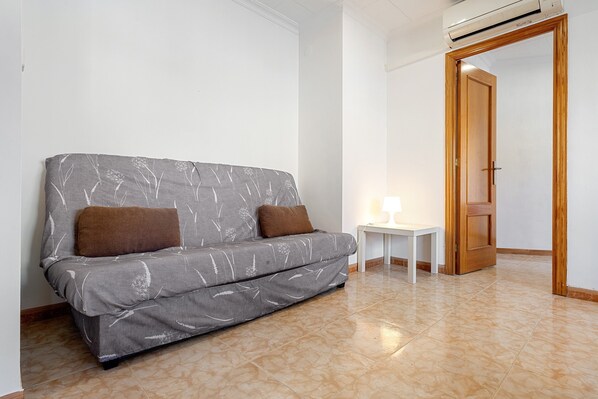 A room with a grey sofa, two brown cushions, a small white table with a lamp, and a wooden door.