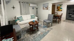 Una sala de estar con un sofá blanco, un sillón gris, una mesa de centro de madera y un comedor con una mesa y sillas de madera.