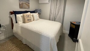 Un dormitorio con cama, mesita de noche, aire acondicionado, ventana con cortinas y un cubo de basura pequeño.