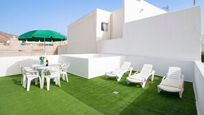 Une terrasse extérieure moderne avec une pelouse verdoyante, des chaises longues blanches, une table avec des chaises et un parasol vert.