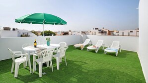 Une terrasse sur le toit, avec un revêtement d’herbe artificielle, des chaises en plastique blanches, une table avec une bouteille et un verre, et un parasol vert.
