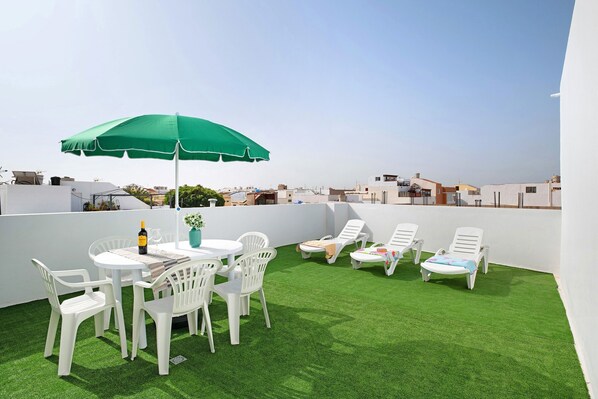 Una terraza en la azotea con césped artificial, sillas de plástico blanco, una mesa con una botella y un vaso, y un paraguas verde.