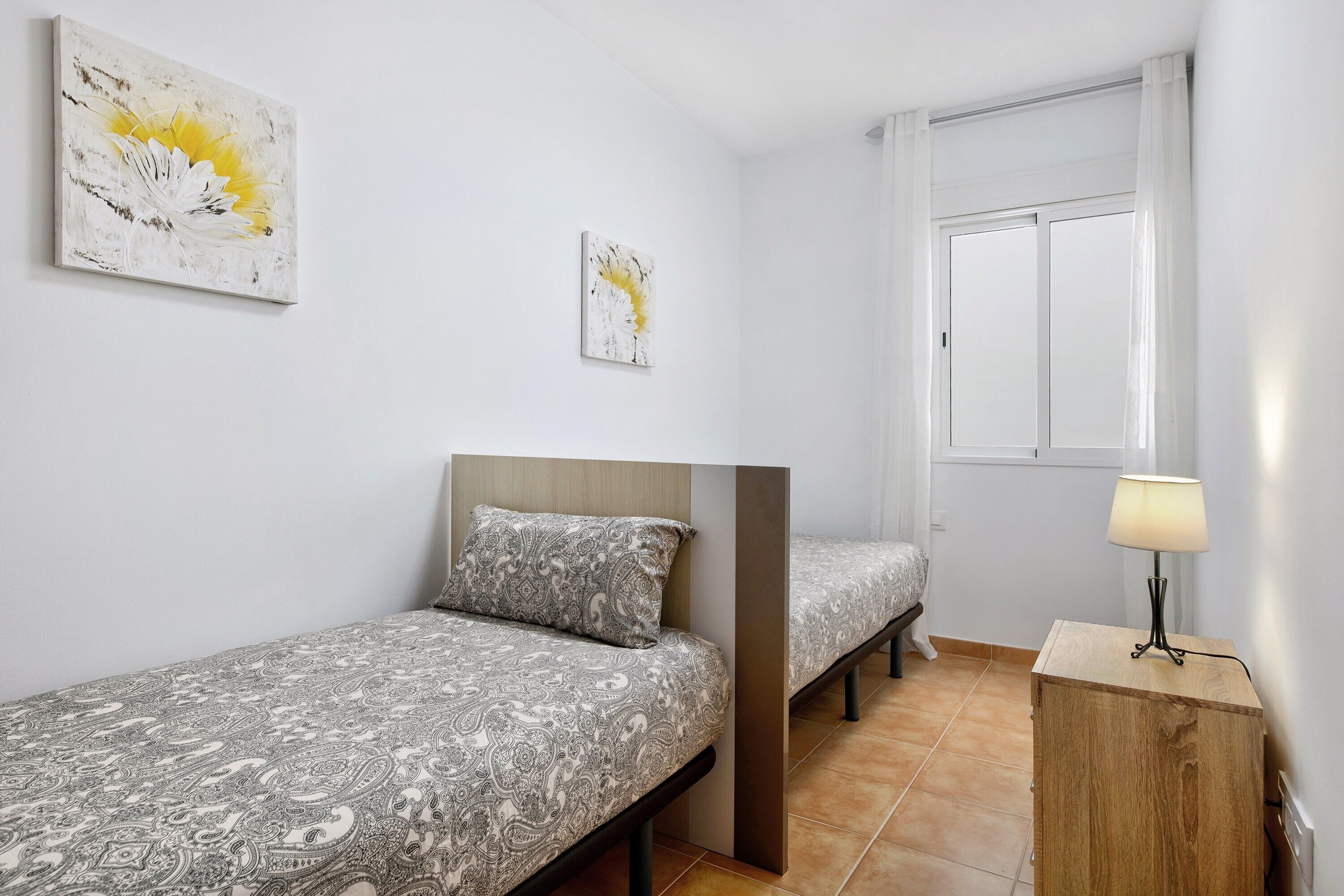 A bedroom with two single beds, a bedside table with a lamp, and two wall-mounted artworks.