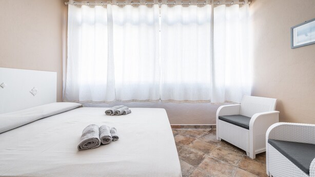 Un dormitorio con una cama, dos toallas enrolladas, una silla blanca y un ventanal con cortinas.