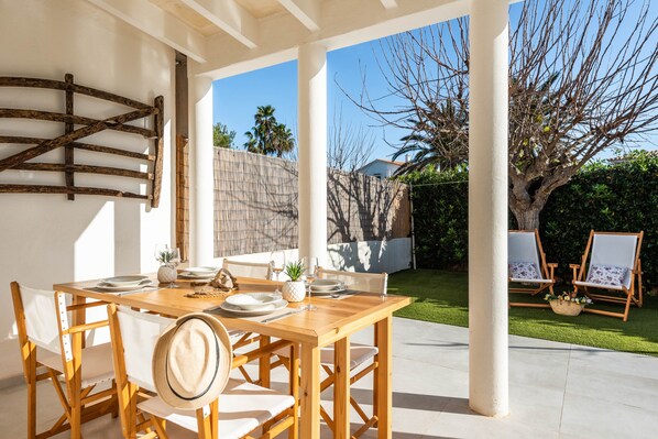 Une terrasse extérieure avec une table en bois dressée pour le repas, entourée de chaises blanches et d’une clôture en bois.