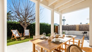 An outdoor patio with a wooden table set for dining, surrounded by white chairs, a large tree, and a view of the sky.
