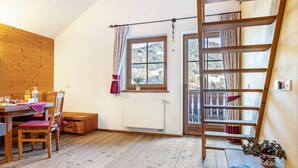 Une pièce avec un plancher en bois, une table à manger avec des chaises, une fenêtre avec des rideaux, un radiateur et une échelle en bois.