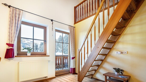 Une pièce avec un plancher en bois, un escalier en bois, une fenêtre donnant sur des arbres couverts de neige et un radiateur.
