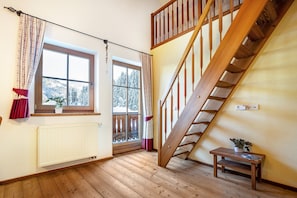 A room with wooden flooring, a wooden staircase, a window with a view of snow-covered trees, and a radiator.