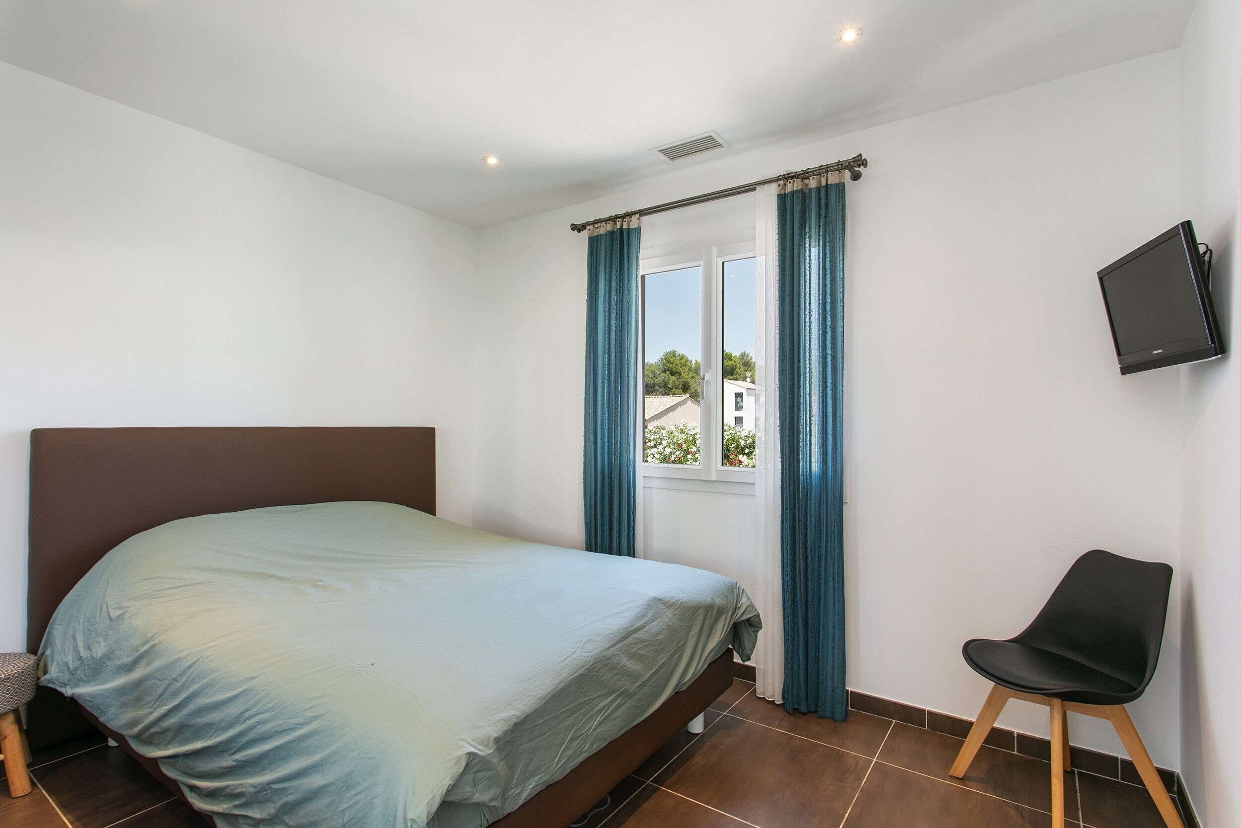 Une chambre à coucher avec un lit, une chaise, un téléviseur et une fenêtre avec des rideaux.