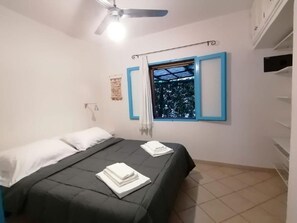 Quarto com cama, ventoinha no teto, janela com persianas azuis e prateleira na parede.