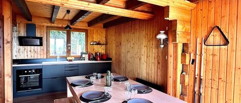 A wooden interior with a dining table set for four, a kitchen with modern appliances, and a large window.