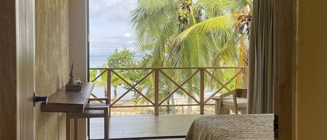 A bedroom with a bed, a small table, a chair, and a view of palm trees and the ocean.